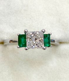 an emerald and diamond ring sitting on top of a white cloth covered surface with two diamonds in the center