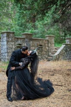 a man and woman dressed in black kissing each other on the ground with trees behind them