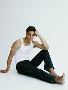 a young man sitting on the floor with his hands behind his head, wearing jeans and a white tank top