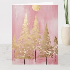 a pink and gold christmas card with trees in the foreground on a marble surface