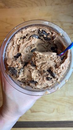 a hand holding a glass with ice cream and chocolate toppings on it, in front of a wooden table