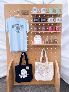 a wooden shelf with two bags and coffee mugs on it next to a t - shirt display