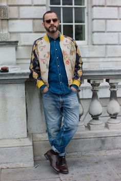 a man leaning against a wall with his hands in his pockets, wearing sunglasses and a colorful jacket