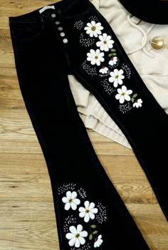 black jeans with white flowers on them sitting on a wooden floor next to a hat