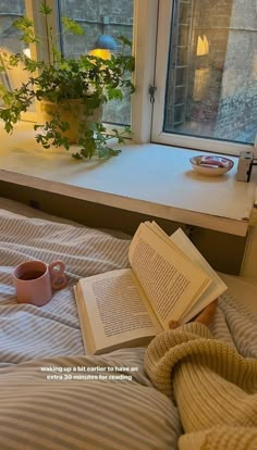 an open book on a bed next to a cup and saucer with a plant