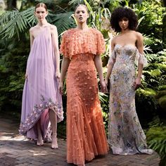 three models in long dresses standing next to each other on a brick walkway surrounded by greenery