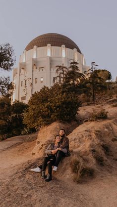 two people sitting on top of a hill next to a large white building with a dome