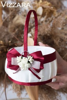 a person holding a white basket with red ribbon and flowers on it, in front of dried plants