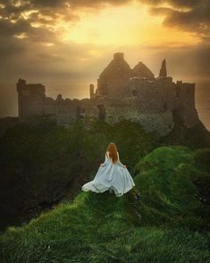 a woman sitting on top of a lush green hillside next to a castle under a cloudy sky