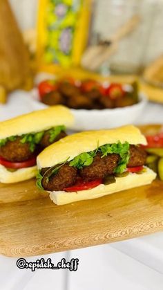 two meatball sub sandwiches on a cutting board with pickles and tomatoes in the background