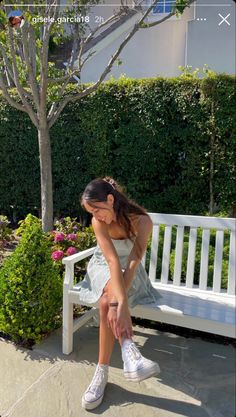 a woman sitting on a white bench in front of a tree and shrubbery with her head down