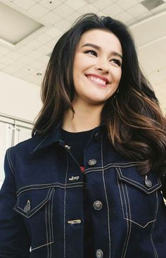 a woman with long hair wearing a denim jacket and smiling at the camera while standing in an office