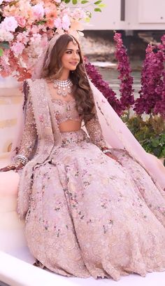 a woman sitting on top of a white bench wearing a pink dress and veil with flowers in the background