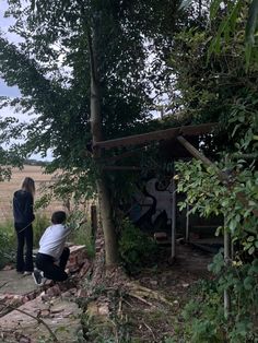 two people are sitting on the ground in front of a tree and some graffiti behind them