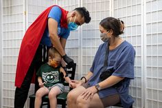 a man in a red cape sitting on a chair next to a little boy who is wearing a face mask