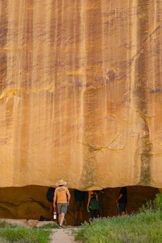 three people are standing at the base of a large rock formation, with grass growing in between them