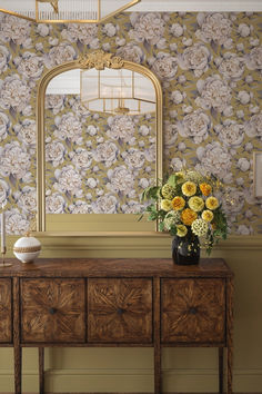 a table with flowers on it in front of a wallpapered mirror and lamp