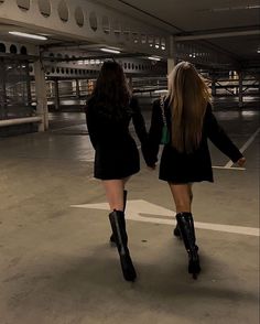 two women in short skirts and black boots walk through an empty parking lot holding hands