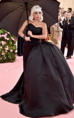 lady in black gown holding an umbrella while standing on pink carpet with other people behind her