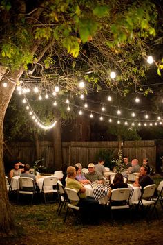 a group of people sitting around a table under string lights in a backyard at night