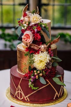 a three tiered cake decorated with flowers and gold leaf accents on a table in front of a window