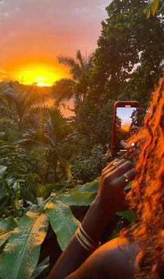 a woman taking a photo with her cell phone in front of the sun setting over some trees