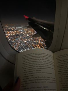 an airplane window with a book in the foreground and city lights seen through it