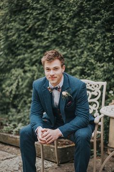 a man in a blue suit sitting on a white chair next to some trees and bushes