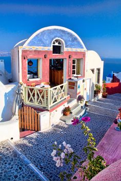 a white and pink building with flowers on the ground in front of blue water behind it