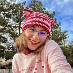 a woman wearing glasses and a knitted cat hat smiles at the camera while sitting on a bench