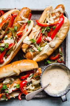 three hot dogs on buns with peppers, onions and cheese next to dipping sauce