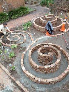 two men are working in the garden with bricks and brickwork around them, while another man is using a wheelbarrow