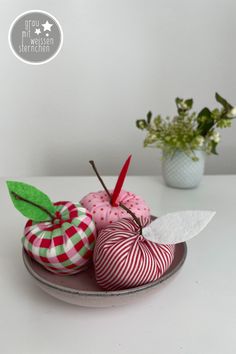 three apples are sitting on a plate next to a vase with green leaves and flowers