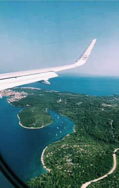 an airplane wing flying over a lush green forest covered island in the middle of the ocean