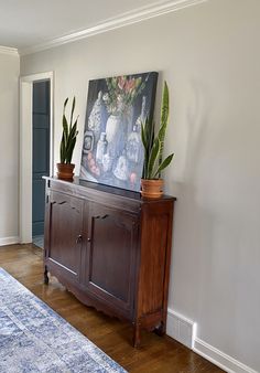 a wooden cabinet with two plants on top of it next to a painting and rug