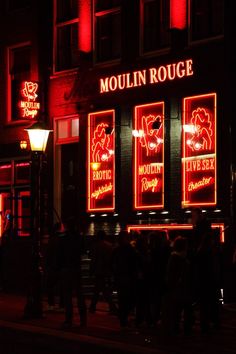 people are walking on the sidewalk in front of a building lit up with red lights