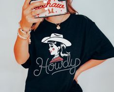 Get ready to turn heads with the "Howdy Cowgirl" graphic tee from Lasso & Lace. This unique design features a retro-inspired cowgirl graphic, complete with her bold lipstick and classic cowboy hat, making it the ultimate statement piece for western lovers. The word "Howdy" is creatively written in a rope-style script, adding a perfect touch of rodeo charm to this tee. Crafted from the super soft and lightweight Bella Canvas 3001, this shirt offers comfort and durability for everyday wear. Whethe Cowgirl Graphic, Vintage Western Shirt, Rodeo Events, Classic Cowboy, Bold Lipstick, Chapeau Cowboy, Cowgirl Aesthetic, Western Graphic Tees, Rodeo Shirts