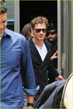 a man in sunglasses is walking out of a building with another man behind him and other people looking on