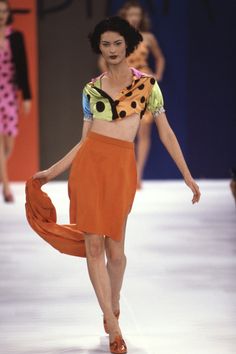 a woman is walking down the runway wearing an orange skirt and polka dot top with her hands in her pockets