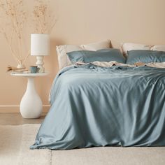 a bed with blue sheets and pillows next to a white vase filled with flowers on a table