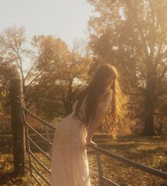 a woman in a white dress leaning on a fence with her hands behind her back