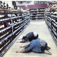 two people laying on the ground in a store aisle with bottles of wine behind them