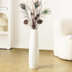 a white vase with feathers and pineconis in it on the floor next to a chair