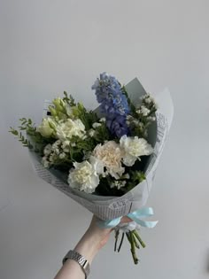 a person holding a bouquet of white and blue flowers in their left hand, against a gray wall