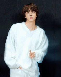 a young man wearing a white sweater standing in front of a black wall with his hands together