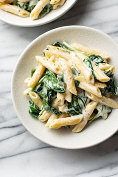 two bowls filled with pasta and spinach on top of a marble countertop next to each other