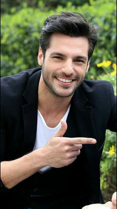 a man pointing at something while sitting in front of some bushes and flowers with his finger up