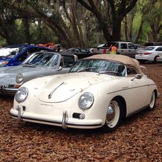 an old white car is parked in the dirt with other cars behind it and trees