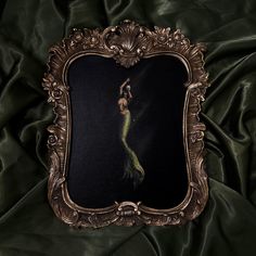 an ornate gold frame with a painting of a mermaid in the middle on a green satin background