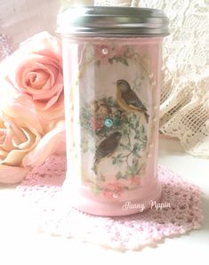 a pink jar with two birds on it next to some flowers and lace doily